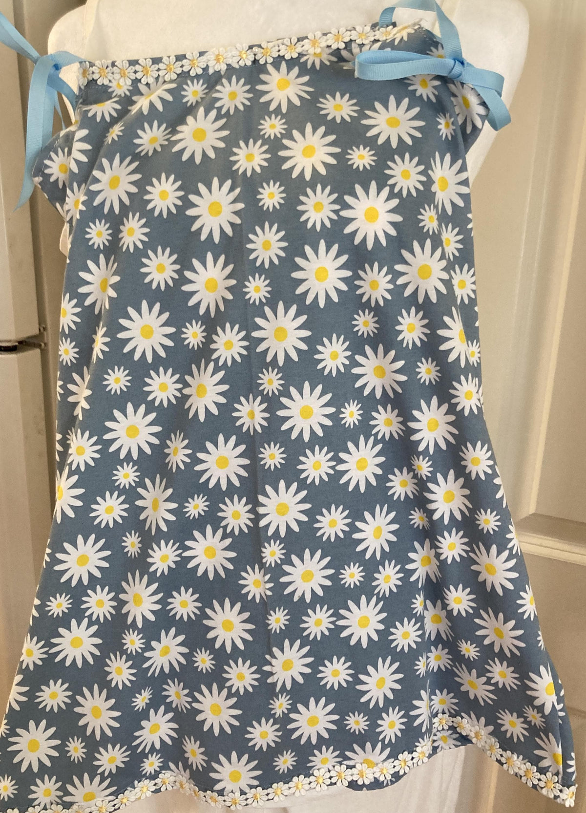 May include: A blue apron with a white daisy pattern and yellow centers. The apron has blue ribbon ties and a white daisy trim along the top and bottom edges. The apron is made of fabric.
