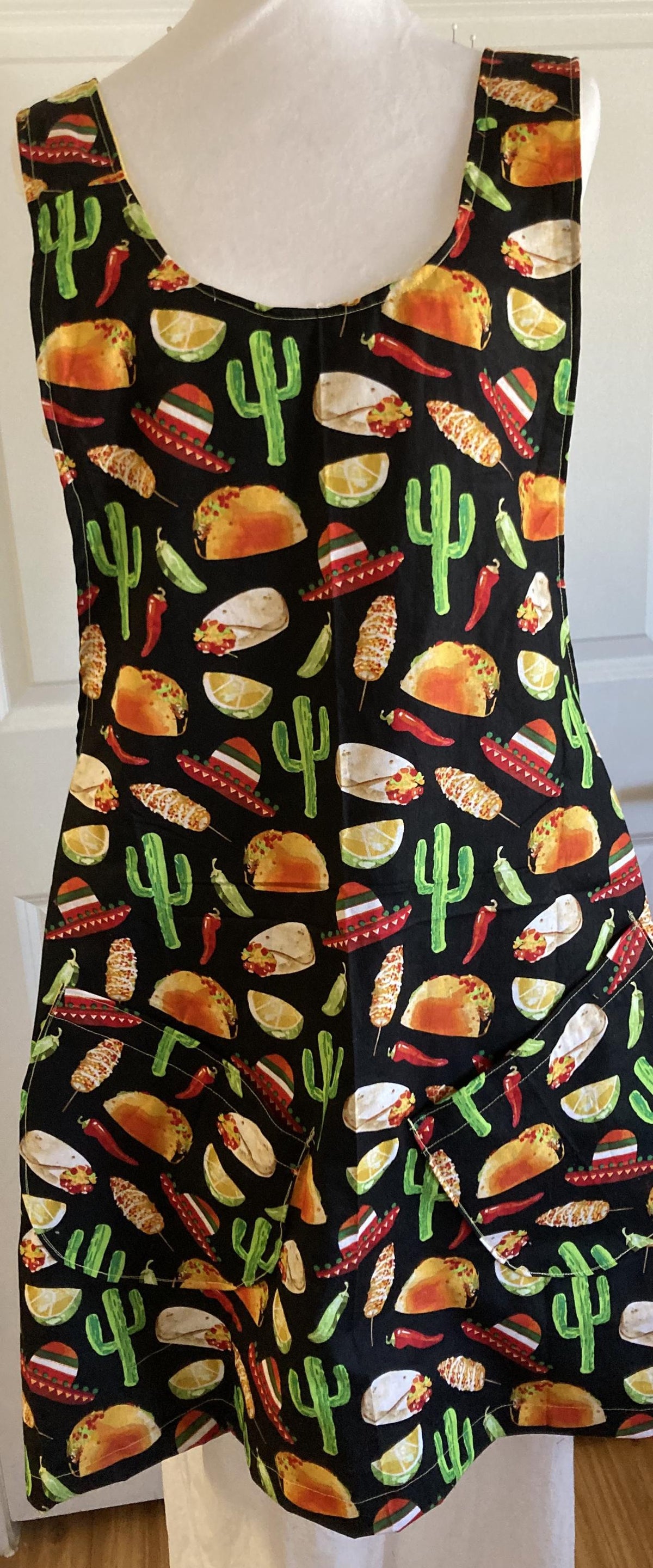 May include: Black apron with a colorful pattern of tacos, sombreros, cacti, limes, and chili peppers. The apron has two pockets and is designed to be worn over the head.