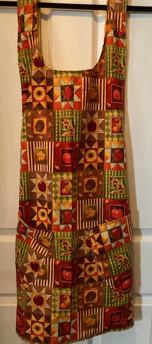May include: A full-length apron featuring a patchwork pattern of autumn-themed squares. The design includes sunflowers, pumpkins, acorns, corn, and leaves in shades of brown, orange, red, yellow, and green. It has two front pockets and shoulder straps.