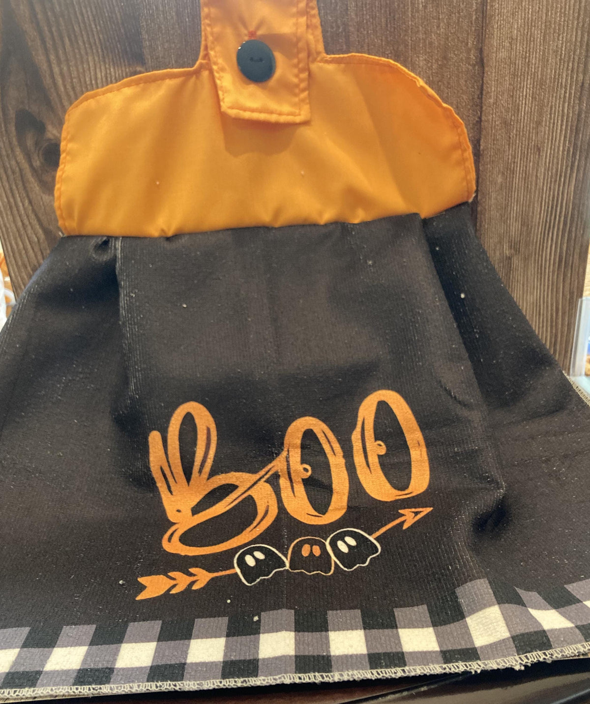 May include: A black hanging kitchen towel featuring an orange top section with a black button closure. The black fabric displays the orange text BOO above an orange arrow graphic with three small ghost illustrations and a grey and black checkered border.